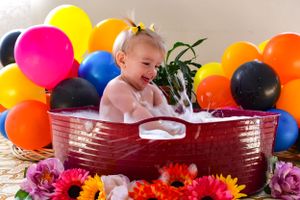 Baby sitting in red basin surrounded by colorful balloons and flowers.