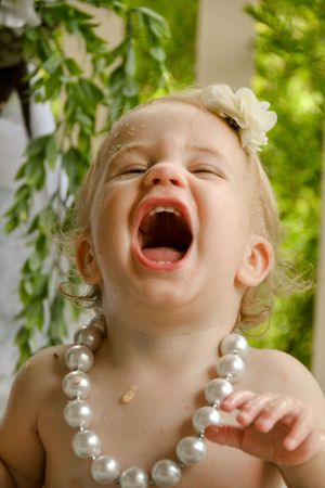 Laughing child wearing oversized pearl necklace against green foliage background.