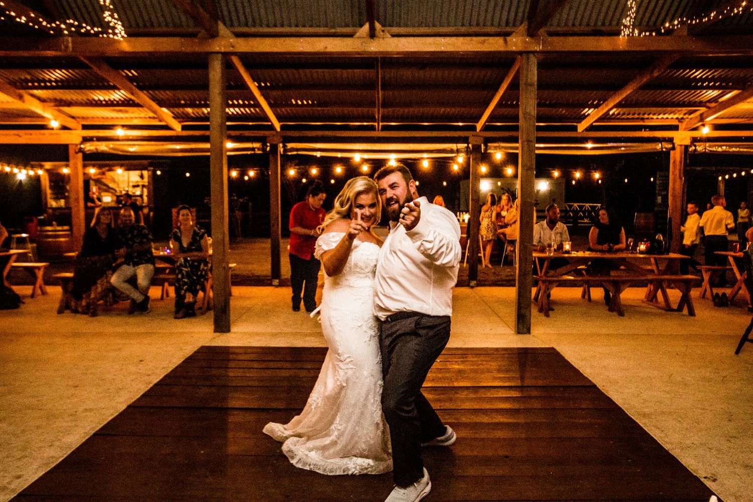 Wedding couple dancing on wooden floor under rustic barn venue with string lights.