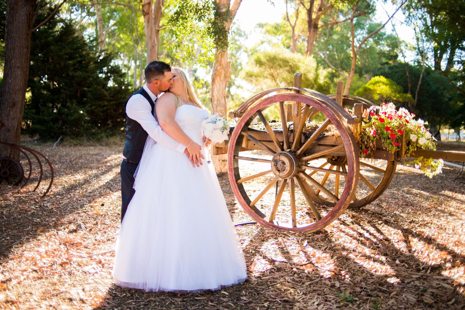 Wedding couple embracing by rustic wooden wagon wheel in dappled sunlight.