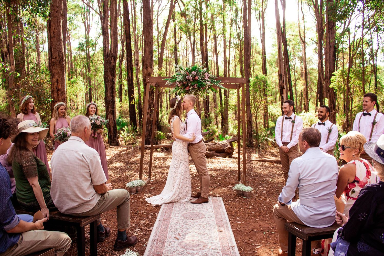 Forest wedding ceremony under wooden arch with guests seated on vintage rugs.