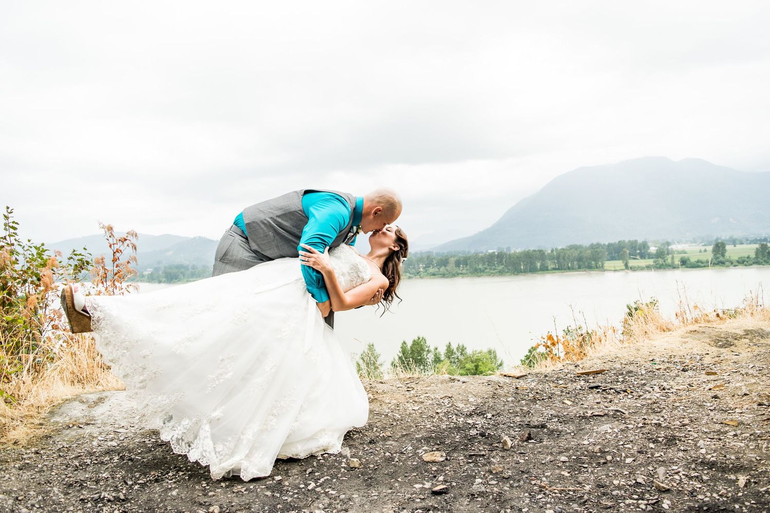 Dynamic dip pose against mountain lake backdrop with flowing wedding dress.