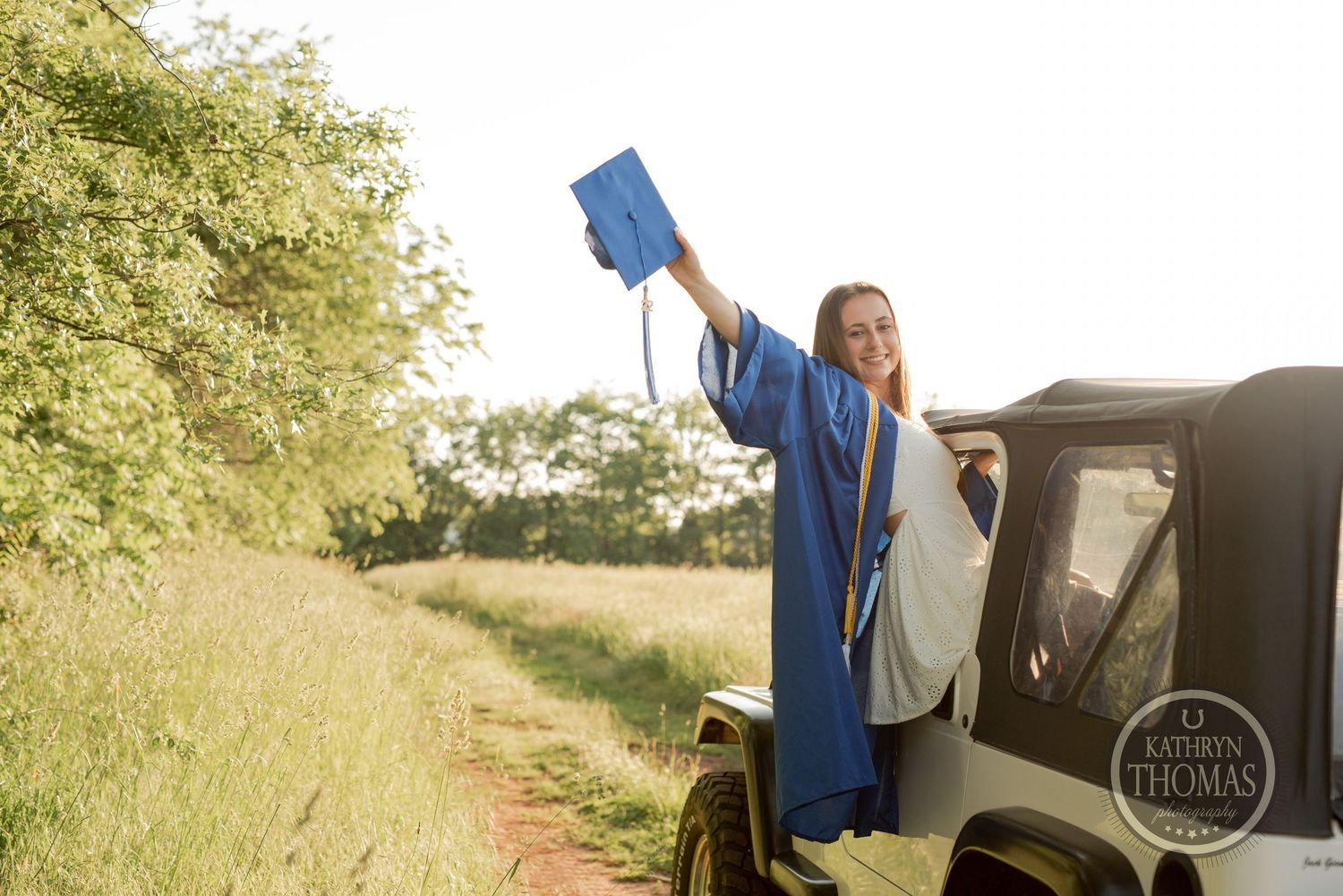 Graduate celebrates by holding cap high while standing in green Jeep on dirt path.