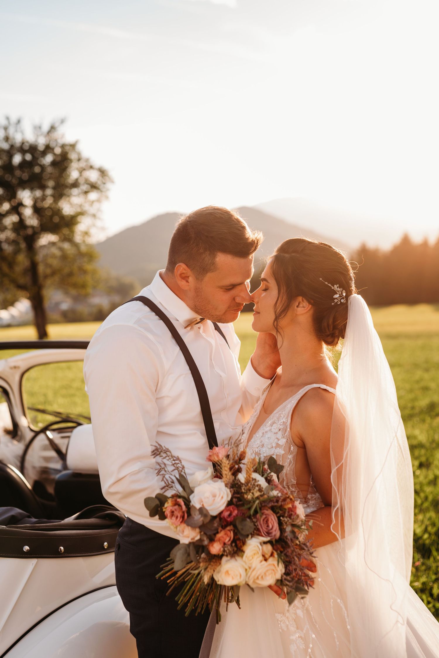 A bride with bouquet and groom share a tender moment at sunset with mountains in background.