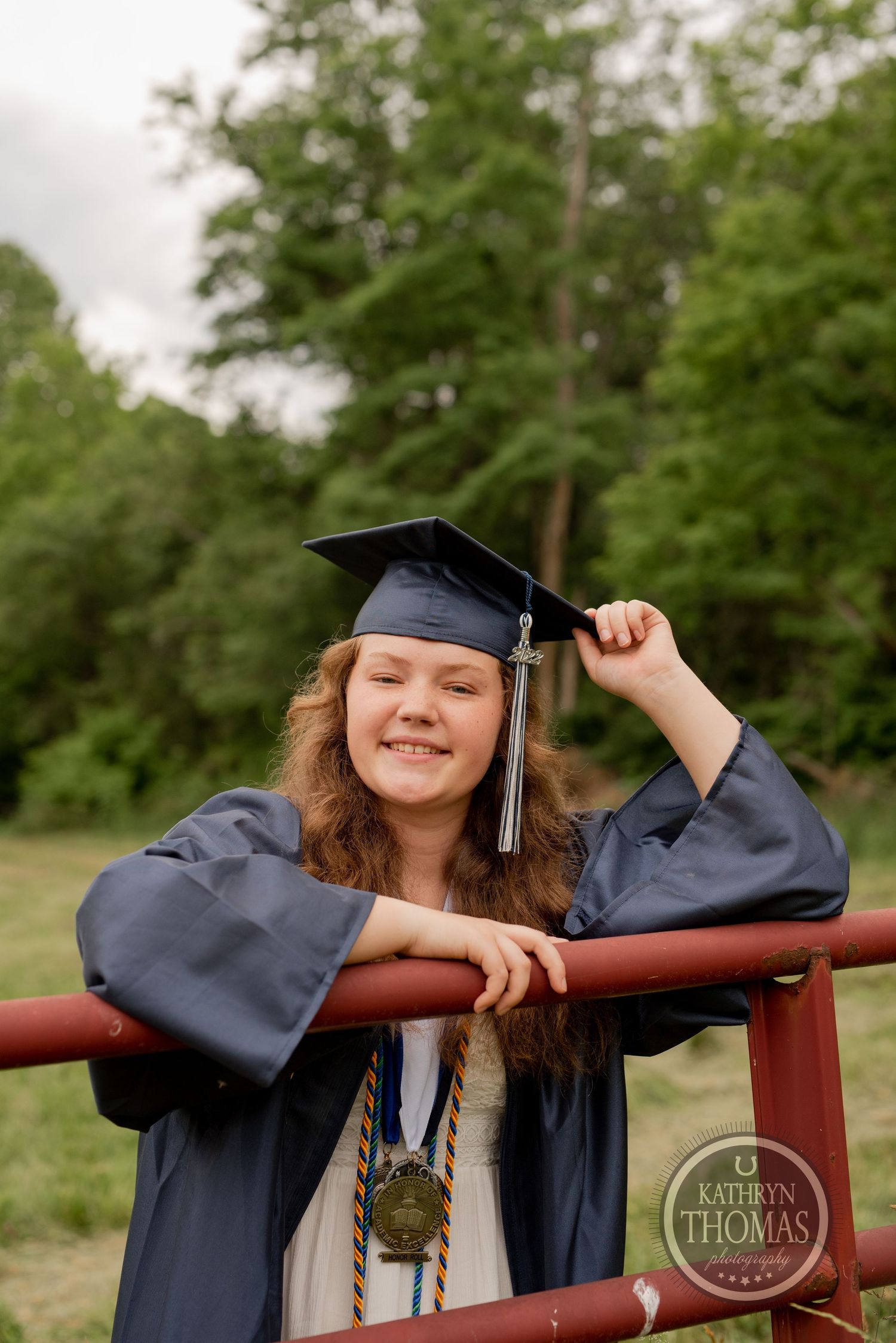 Graduate in academic regalia leans on red railing outdoors with trees in background.