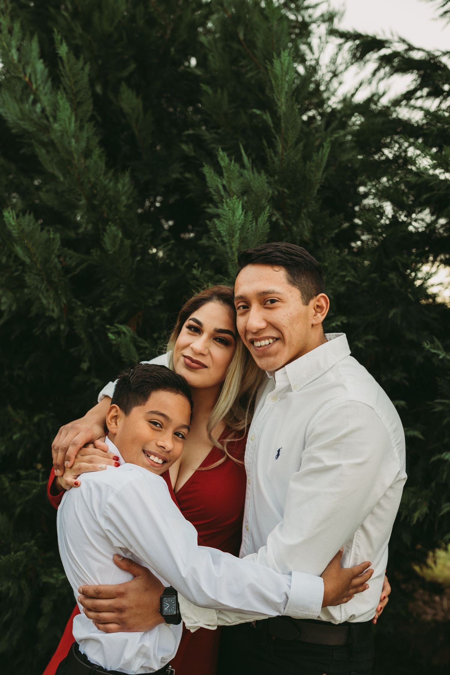 A warm family portrait outdoors showing three people embracing against a backdrop of evergreen trees at sunset.