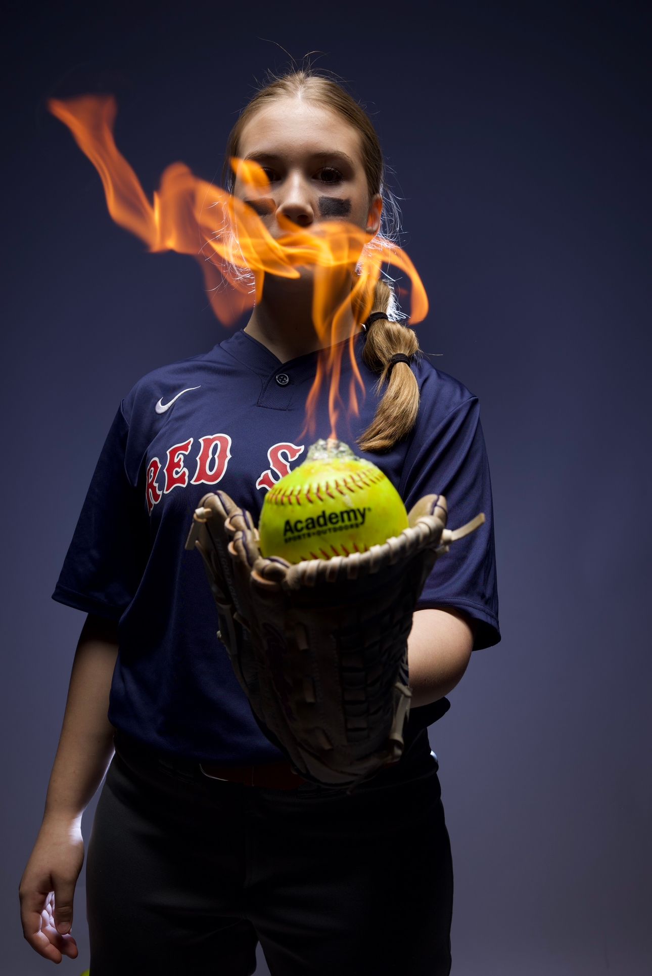 Softball player in Red Sox jersey with flaming softball in glove against dark background.