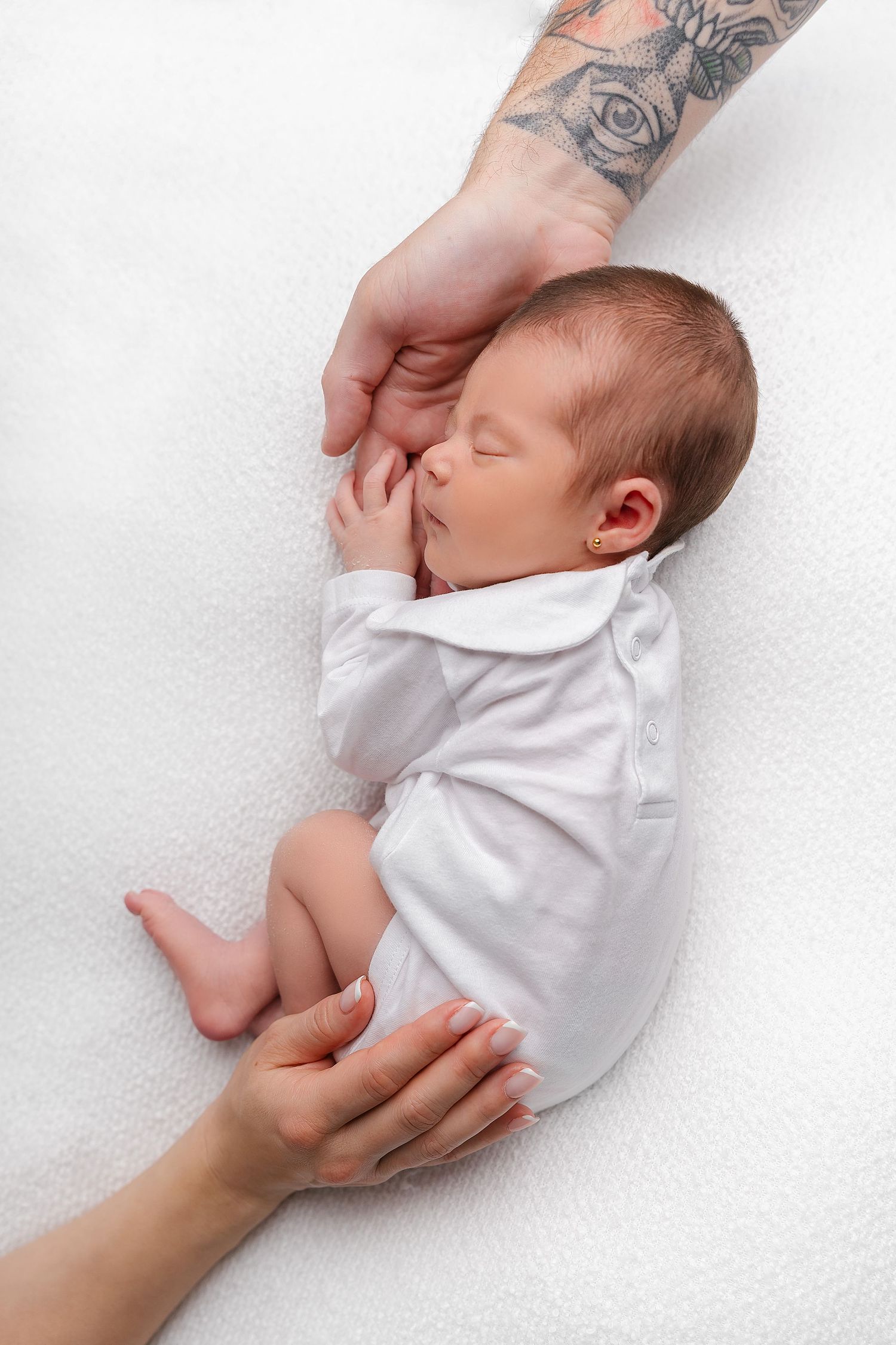 A sleeping newborn baby in white clothing lies on textured white fabric with hands gently supporting them.