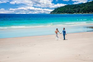 Scenic beach landscape with turquoise waters, white sand, and lush green mountains under puffy clouds.