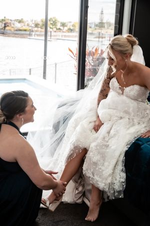 A bride in a white lace dress sitting by a window with natural light streaming in.
