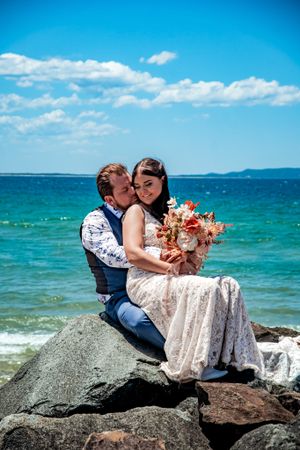 Couple sitting on coastal rocks with turquoise ocean and blue sky backdrop.