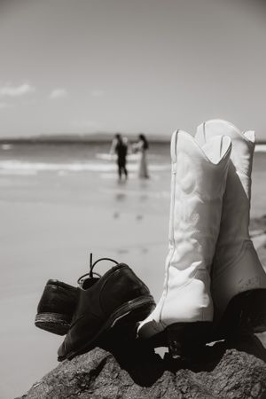 Black and white cowboy boots arranged on rocks at the beach with ocean waves in the background.