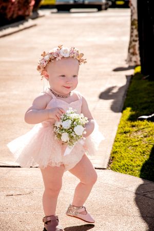 Baby in pink dress and floral crown walking on sunny sidewalk holding flowers.