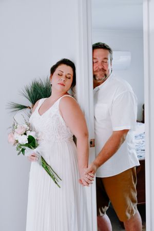 Couple in white attire sharing intimate moment while holding wedding bouquet.