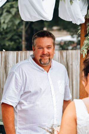 Man in white button-down shirt smiles while seated outdoors with wooden fence in background.