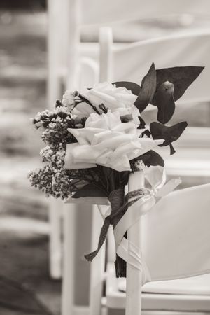 Black and white photo of a delicate floral bouquet tied with ribbon to a white chair at a wedding ceremony.