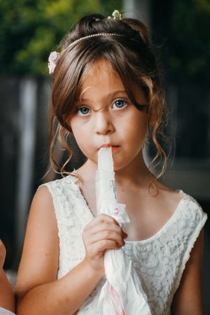 Young child in white lace dress enjoying a frozen treat on a warm day.
