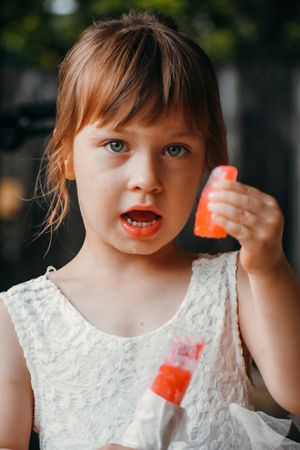Child in white dress holds orange popsicle while making playful expressions.
