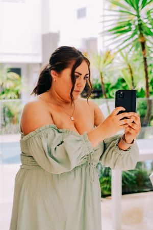 Woman in sage green off-shoulder dress taking a selfie in a tropical setting.