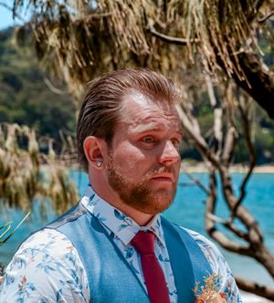Groom in blue vest and red tie smiling during outdoor beach wedding ceremony.