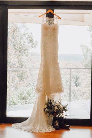 An elegant white lace wedding dress hanging by a window with a floral bouquet below.