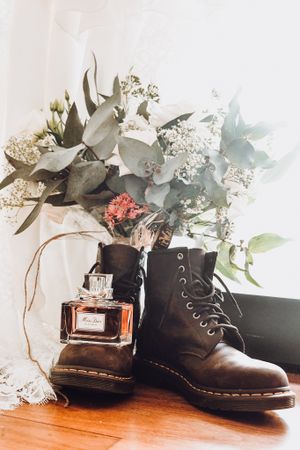 Still life with combat boots, perfume bottle and delicate floral arrangement on wooden surface.