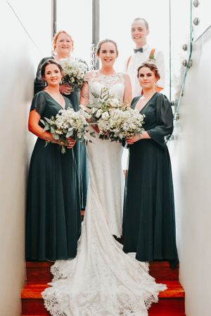 Bridal party in emerald green dresses posing with bride in white lace gown on stairs.