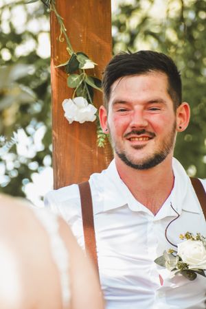 Wedding guest in white shirt and suspenders smiling at outdoor reception with floral decorations.