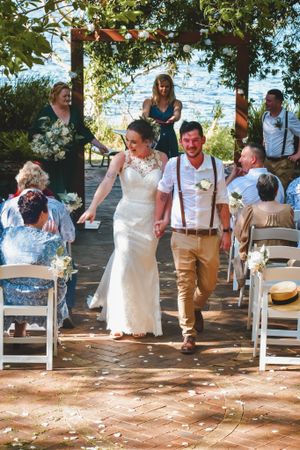 Wedding couple walking down the aisle after ceremony under floral arch with guests seated on white chairs.