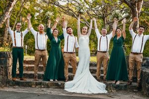 Wedding party in green dresses and khaki suspenders poses joyfully on outdoor steps.