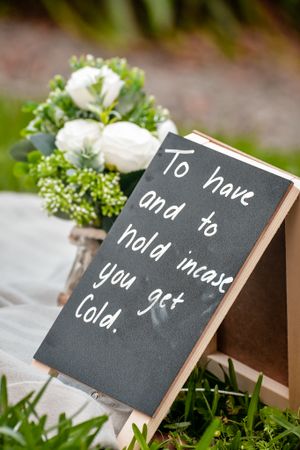 Wedding sign with white flowers reading 'To have and to hold incase you get cold' on chalkboard.