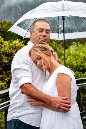 Couple embracing in the rain under a white umbrella on their wedding day.