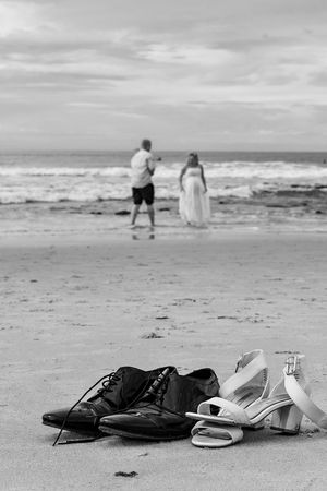 Wedding shoes left on a beach with a couple walking near the ocean waves in black and white.