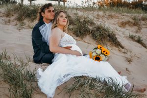 A romantic beach wedding photoshoot featuring a white dress and sunflowers against sand dunes at sunset.