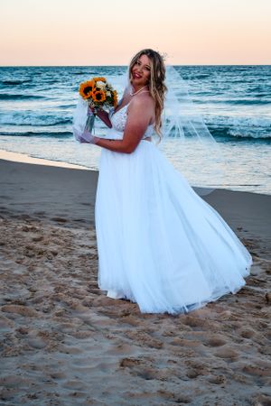 A flowing white wedding dress twirls on a sandy beach at sunset with ocean waves in the background.