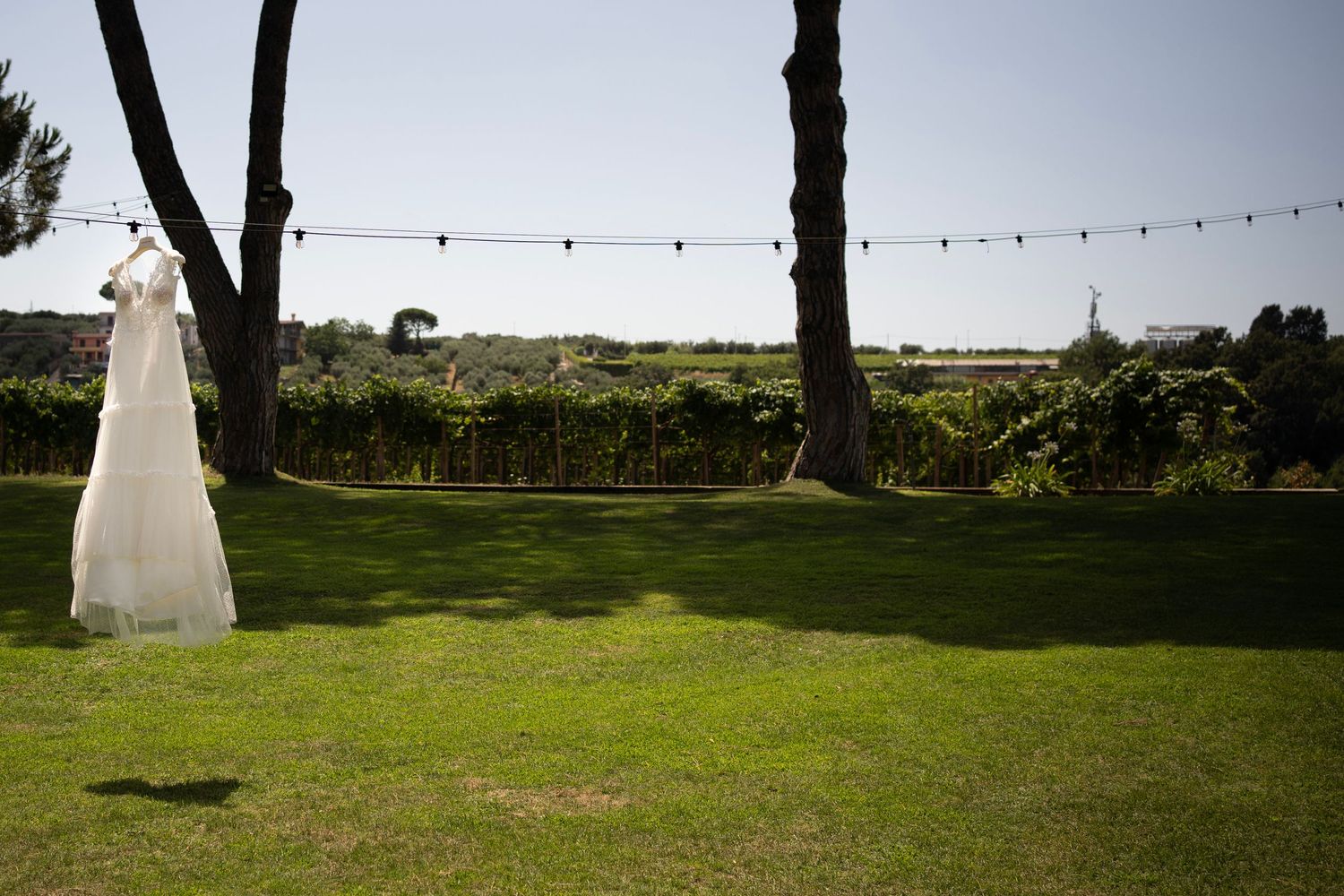 White wedding dress hanging between trees with string lights in outdoor garden setting.