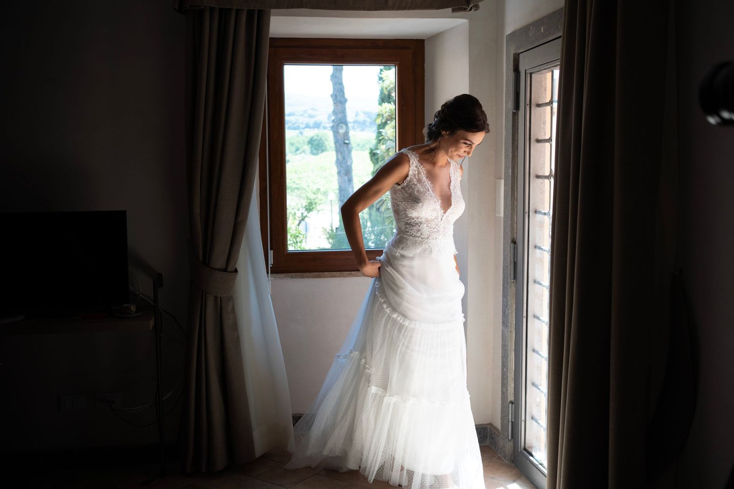 Bridal gown silhouetted against window light in rustic room.