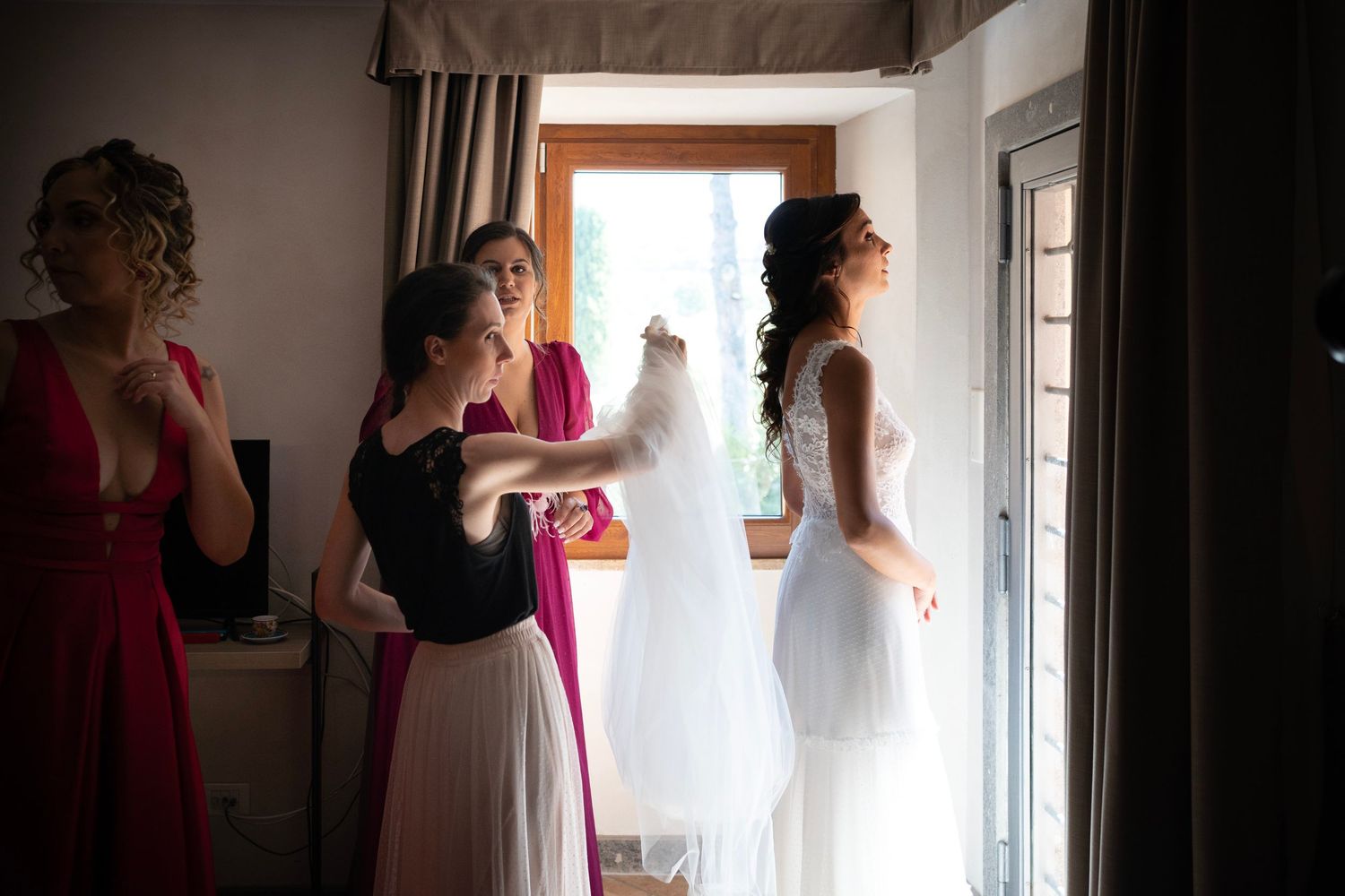 Wedding party members gather around a white dress near a window in preparation for the ceremony.