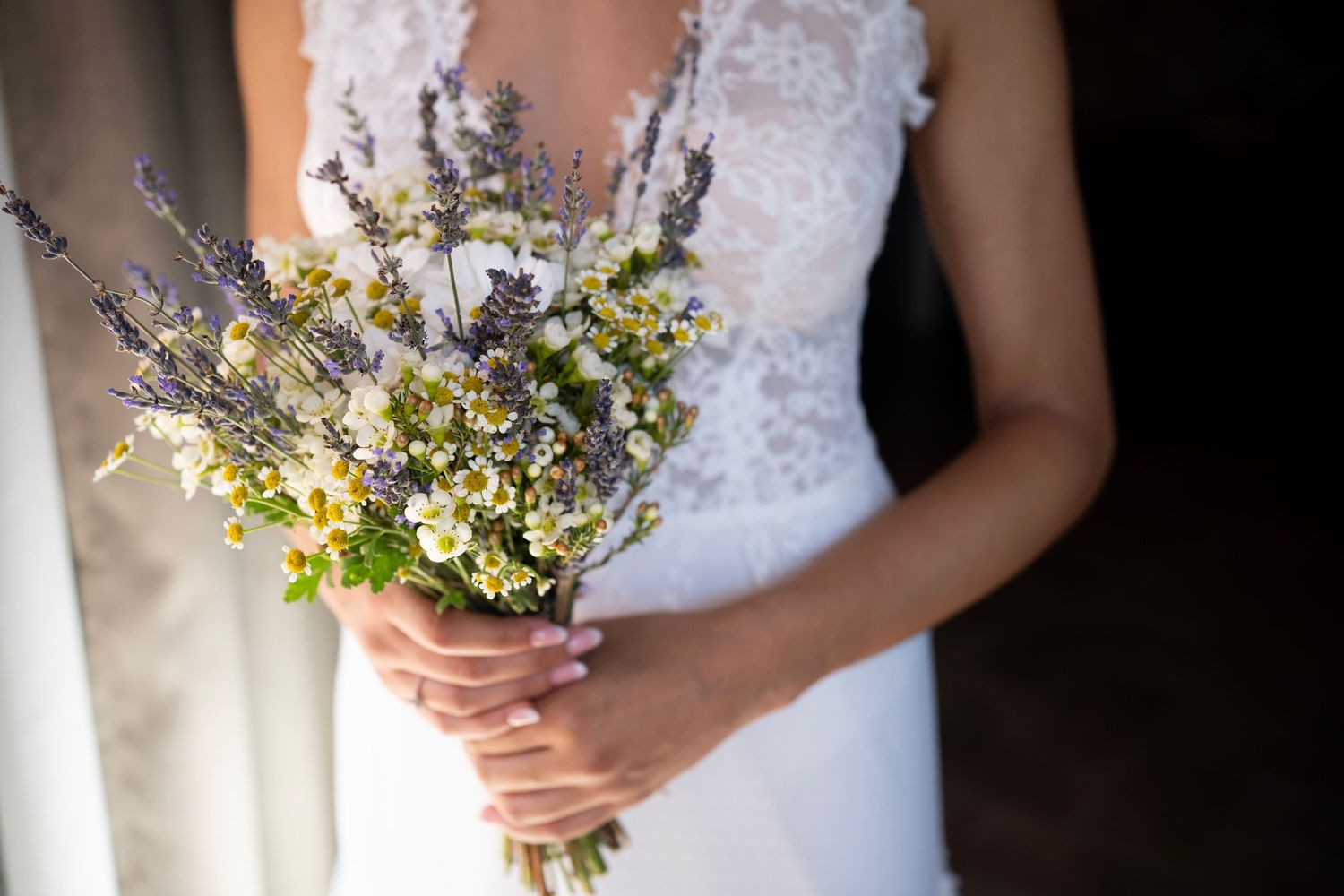 Close-up of a delicate wildflower and lavender wedding bouquet held against a white lace dress.