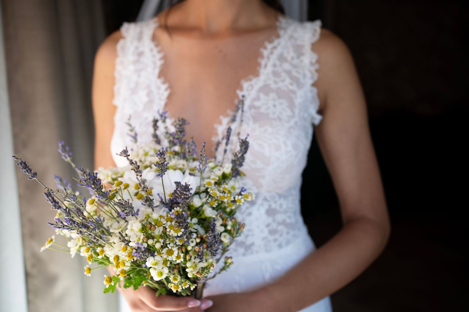 Delicate bouquet of lavender and wildflowers held against a white lace wedding dress.