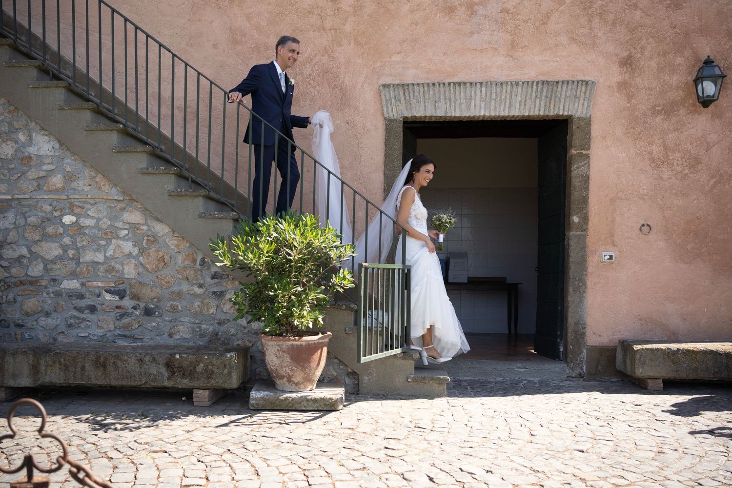 Newlyweds pose on rustic stone staircase against terracotta walls at Mediterranean venue.