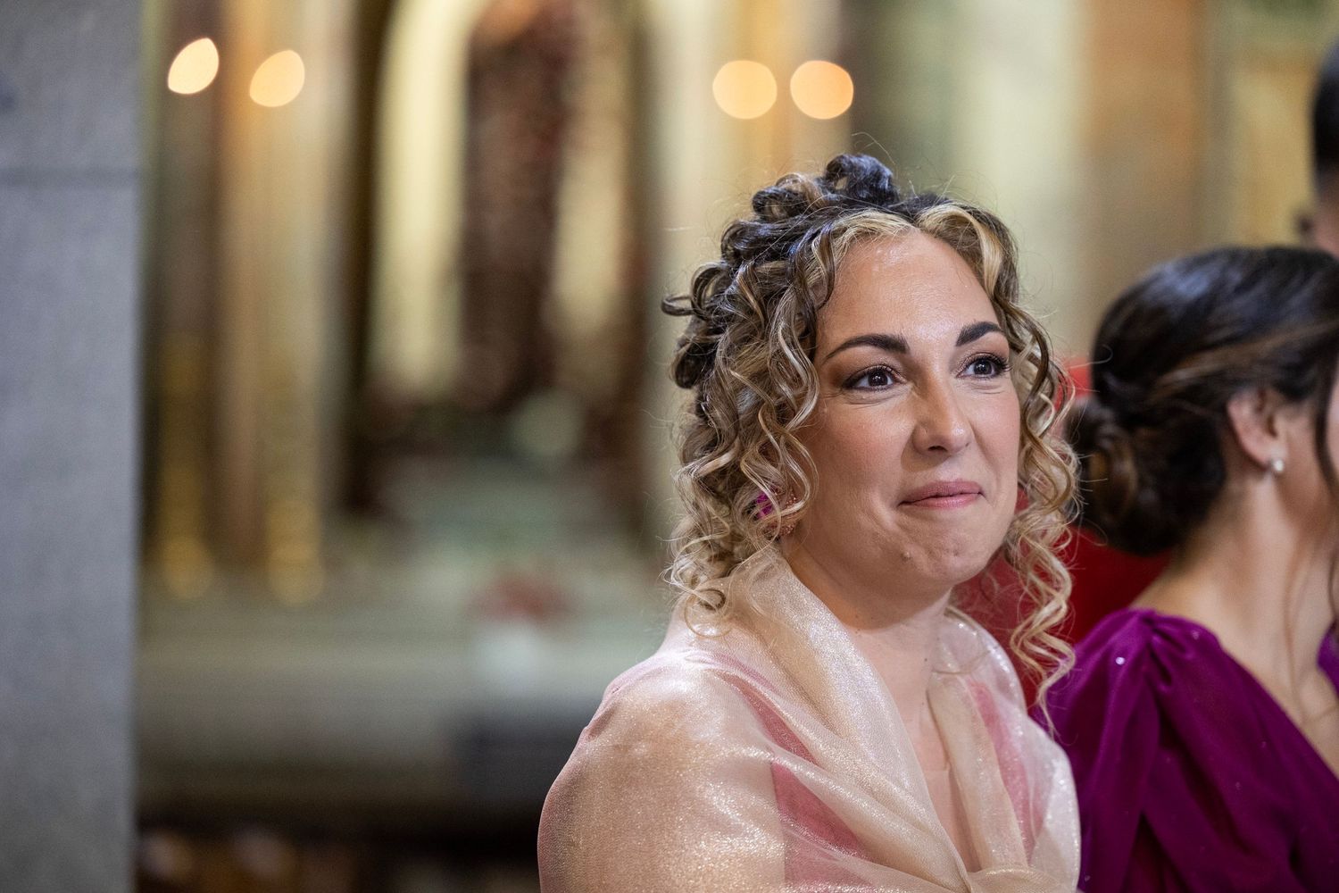 Wedding guest in pink attire smiles during celebration with soft bokeh lights in background.