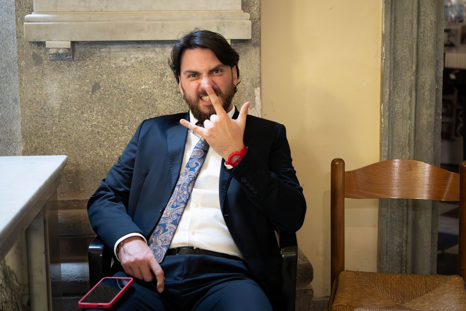 Wedding guest in suit and tie gestures playfully while seated outdoors.