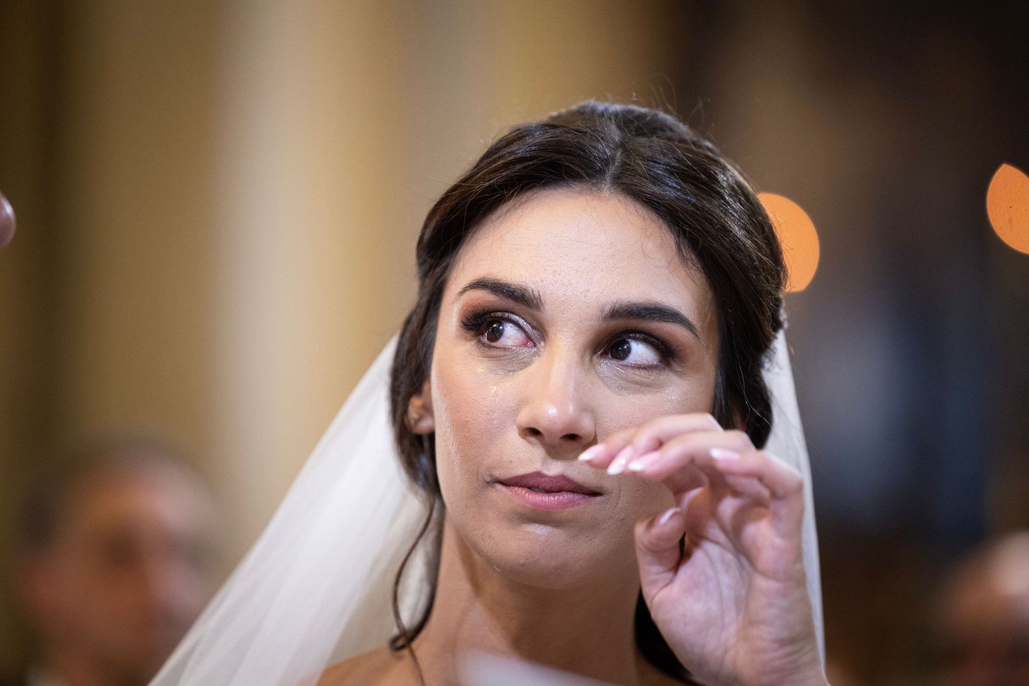 Bride in white veil and elegant makeup gazes upward during emotional ceremony moment.