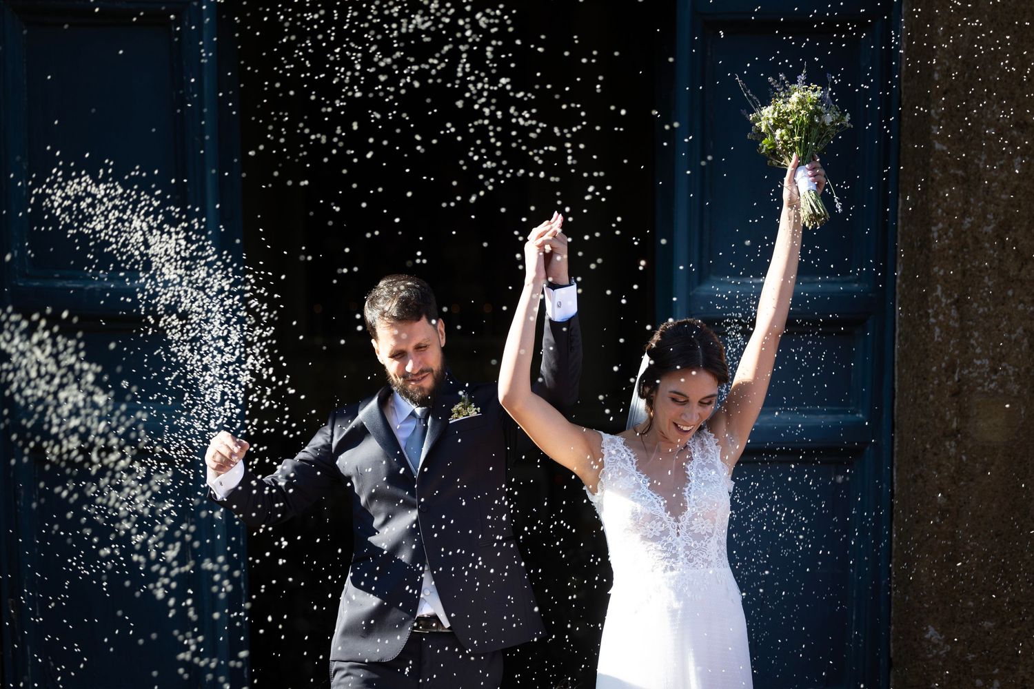 Wedding couple celebrates with upraised arms amid falling confetti against blue doors.