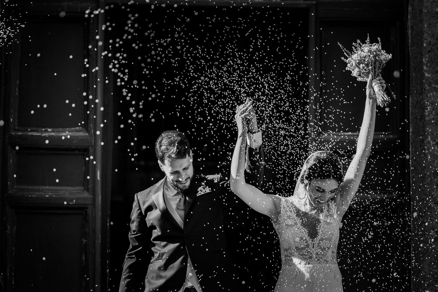 Newlywed couple celebrating under a shower of confetti in dramatic black and white.