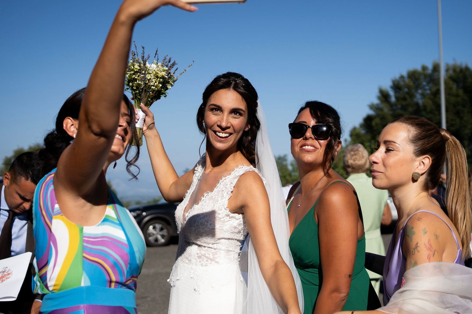 Wedding party celebrates outdoors with bride in white lace dress raising bouquet.