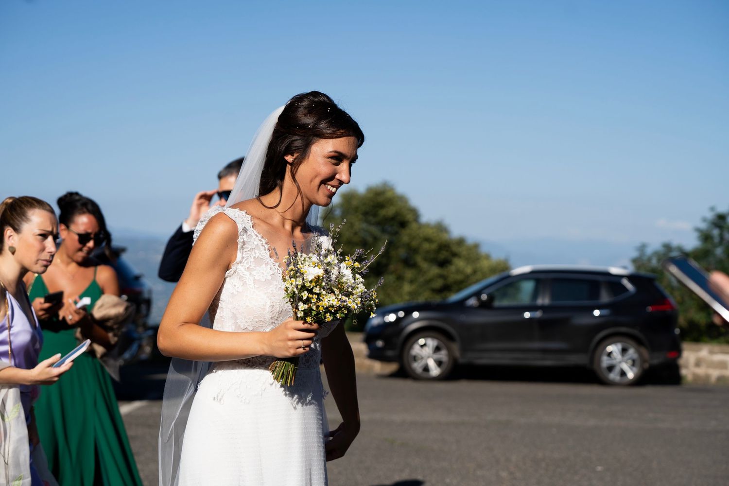 A bride in a white lace gown holds a small bouquet while standing in a sunny outdoor setting.