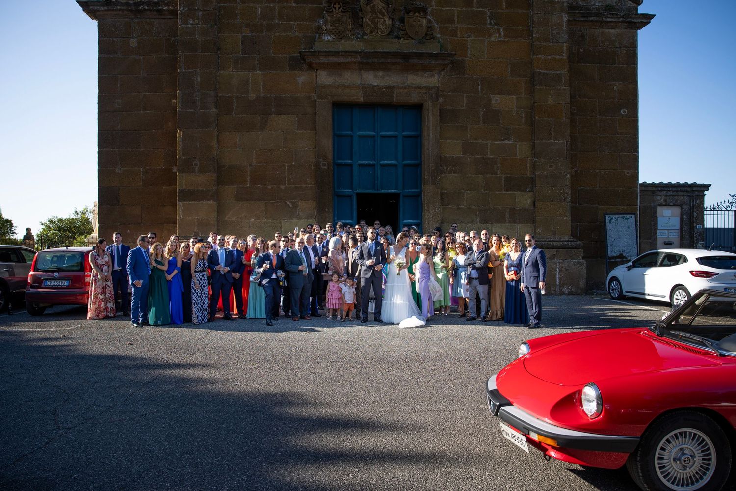 Large wedding group photo outside historic stone building with classic red car in foreground.