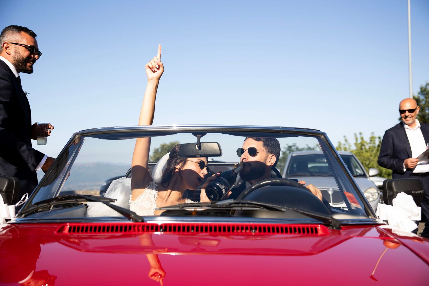 Wedding party celebrates in a red convertible car under bright blue sky.